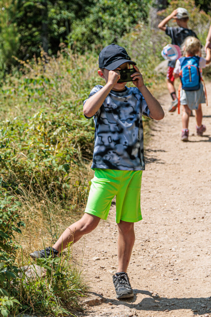 A child wearing a black cap and neon green shorts uses binoculars on a dirt path, while other children walk in the background.  Camp Snap Discount Code