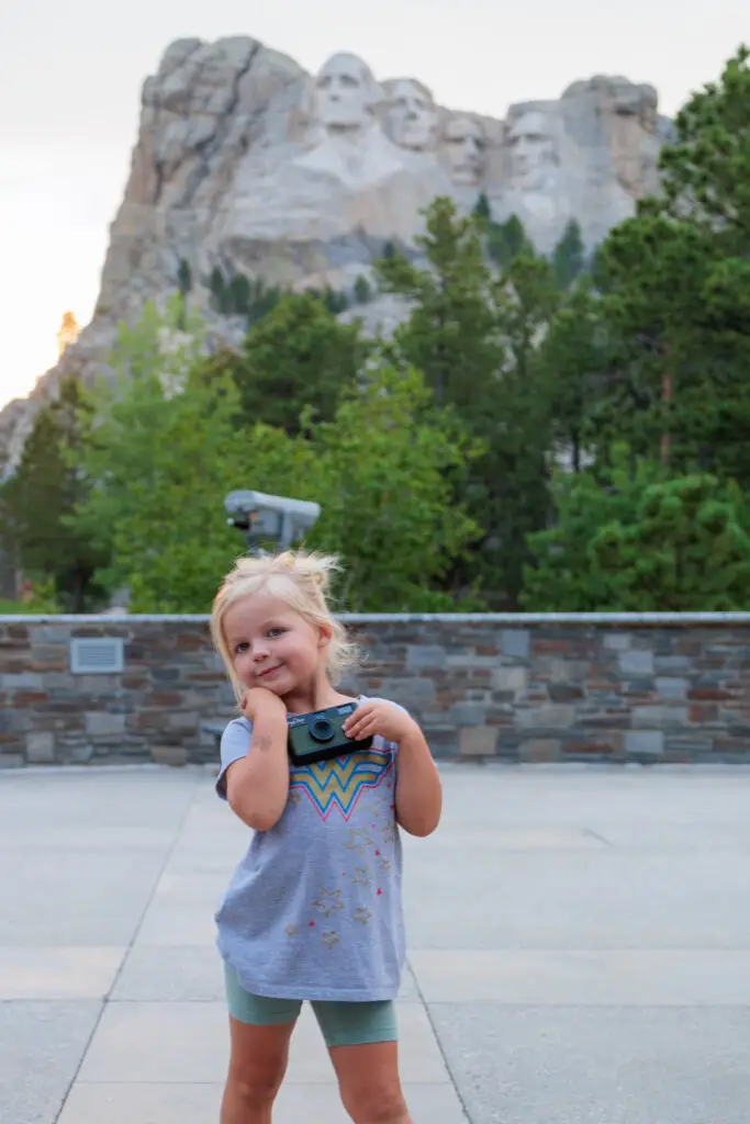 A young girl holds a camera, posing with Mount Rushmore in the background on a clear day.  Camp Snap Discount Code