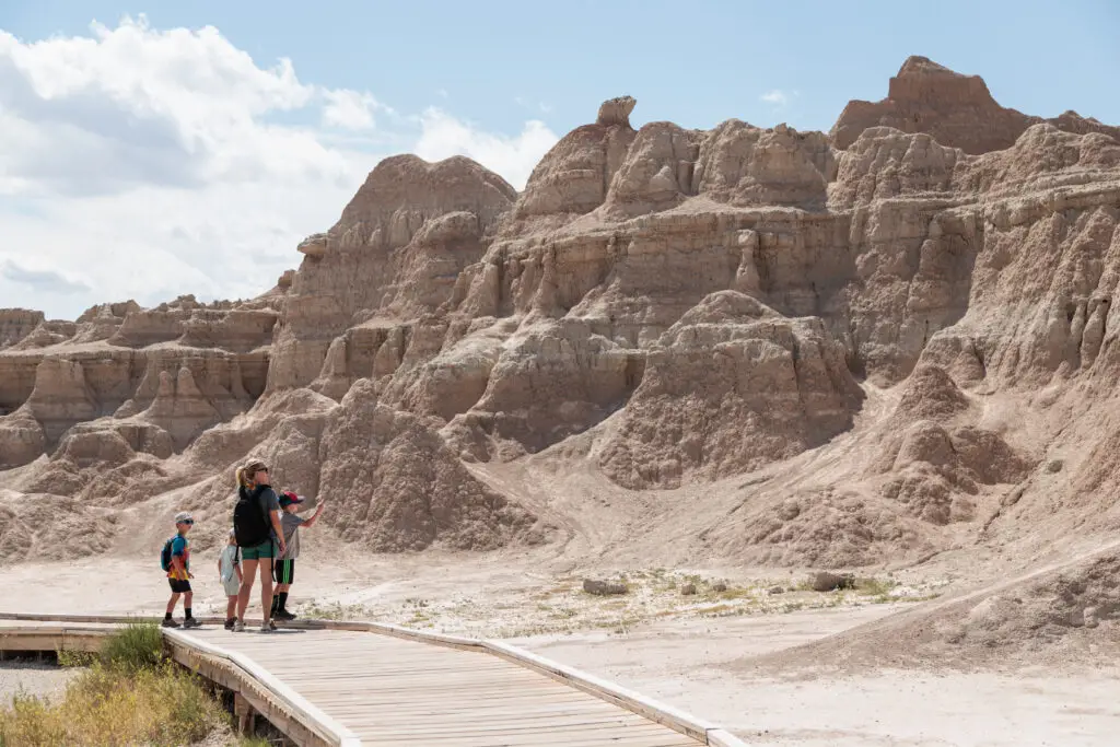 A group of people stand on a boardwalk, observing rocky formations under a clear sky.  Badlands with kids.