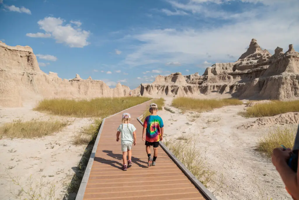 Two children walk on a wooden path through a rocky, desert-like landscape under a clear blue sky.  Badlands with kids.