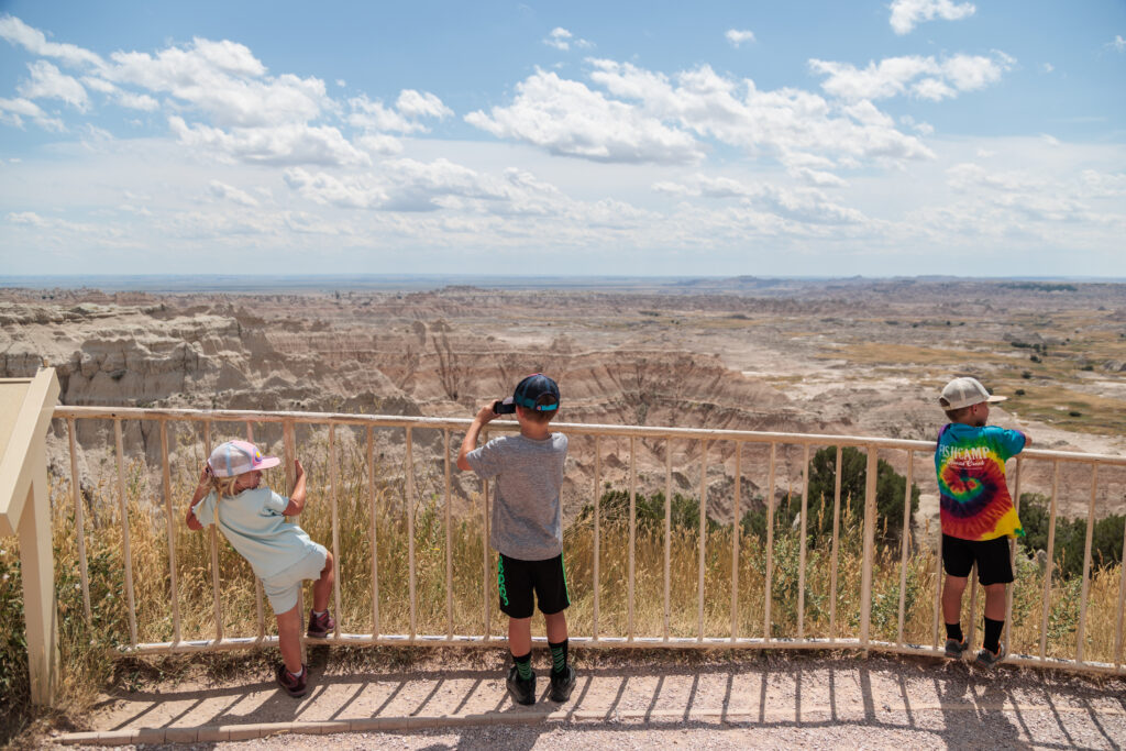 Three children stand at a fence overlooking a vast, rocky landscape on a sunny day. One child wears a tie-dye shirt.  Badlands with kids.