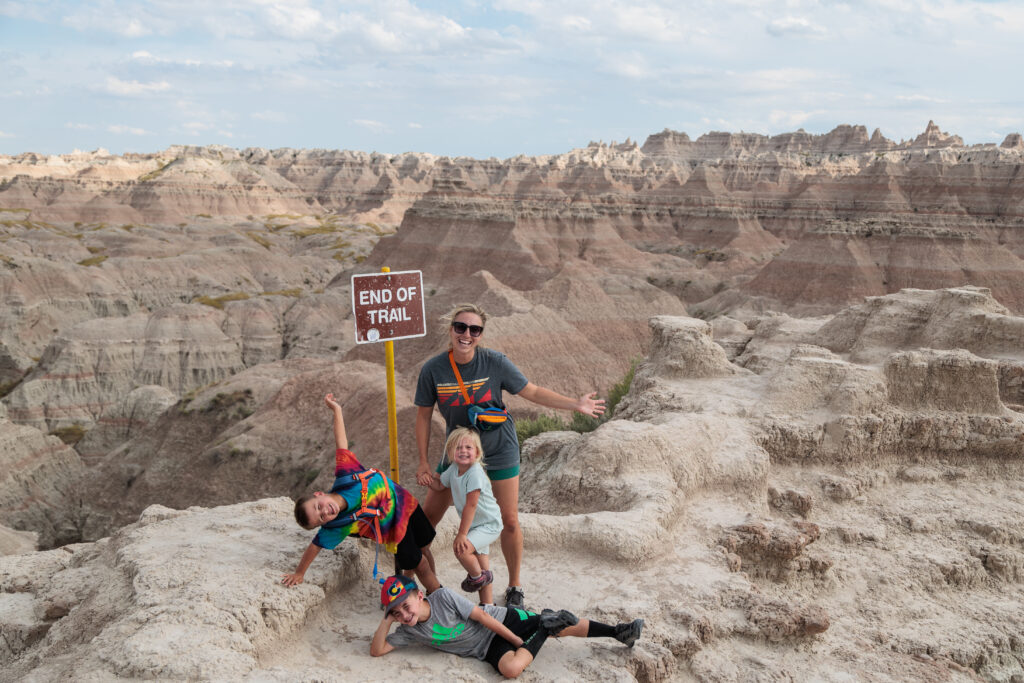 A family poses playfully by an "End of Trail" sign in a rocky desert landscape with layered geological formations under a partly cloudy sky.  Badlands with kids.