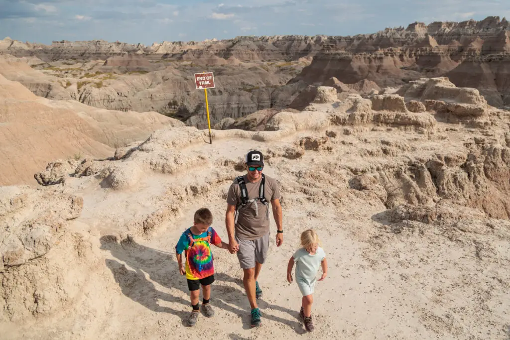 A man walking with two children along a rocky trail in a desert landscape, with a sign indicating the end of the trail in the background.  Badlands with kids.