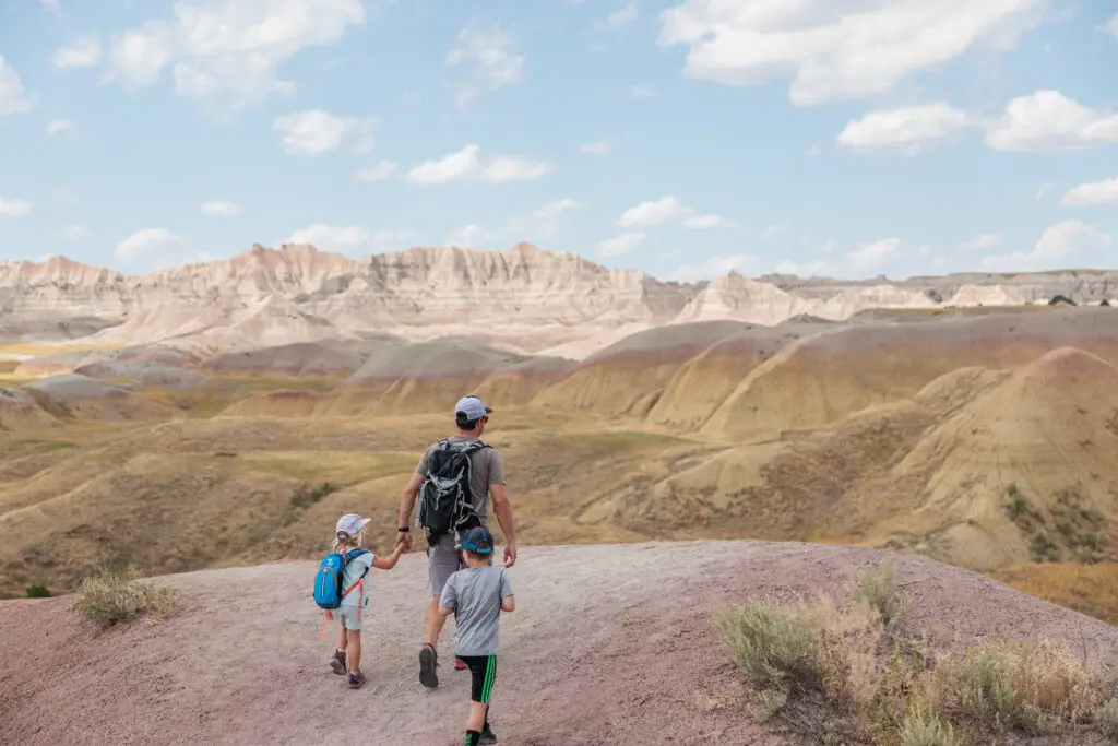 A person with two children hiking on a trail overlooking colorful hills under a partly cloudy sky. Badlands with kids.