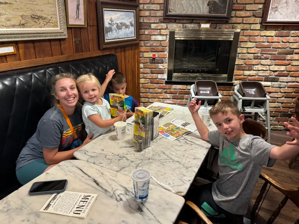 A woman and three children sit at a marble table in a restaurant, with menus and drinks. The background features brick walls and framed pictures. Wall Drug with kids