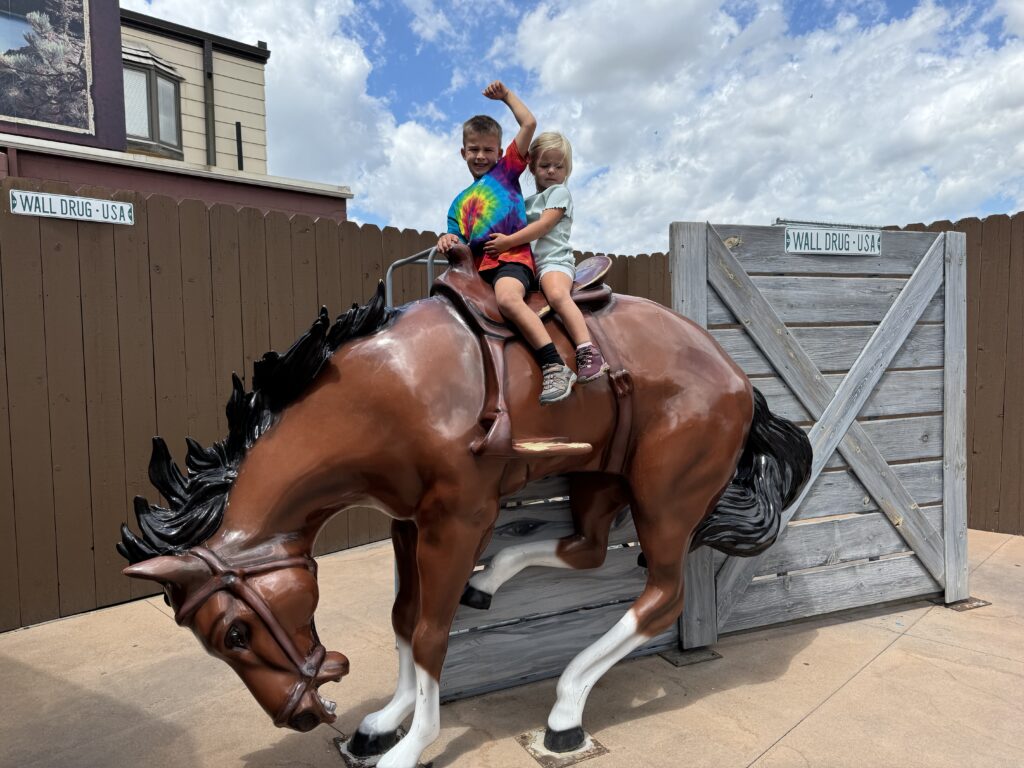 Two children sit on a mechanical horse in front of a brown fence. The boy wears a tie-dye shirt and raises his arm. The girl wears a light blue shirt and holds onto the saddle. Wall Drug with kids