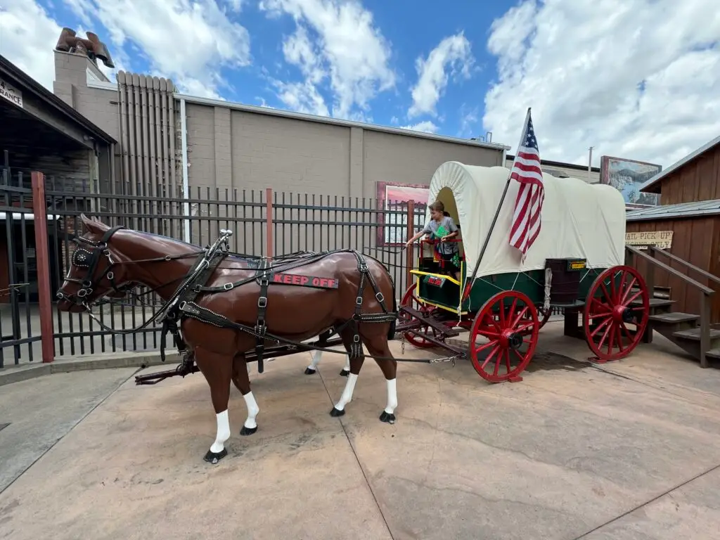 A covered wagon with red wheels hitched to two model horses is displayed outside a building. An American flag is attached to the wagon, and a figure sits inside.  Wall Drug with kids