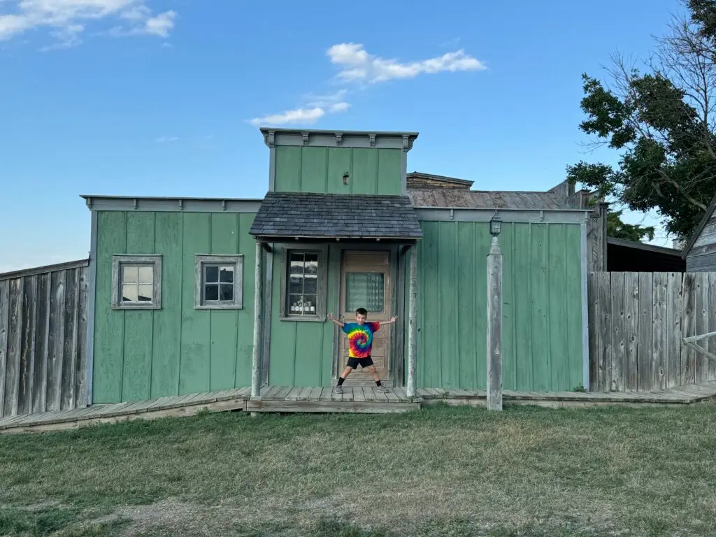 Child in a tie-dye shirt stands with arms outstretched in front of a green wooden building on a clear day. 1880 Town with kids