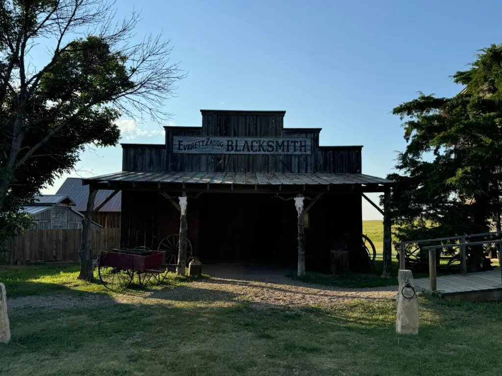 A rustic blacksmith shop with a wooden facade, surrounded by trees. A red cart sits alongside the building under a clear blue sky. 1880 Town with kids