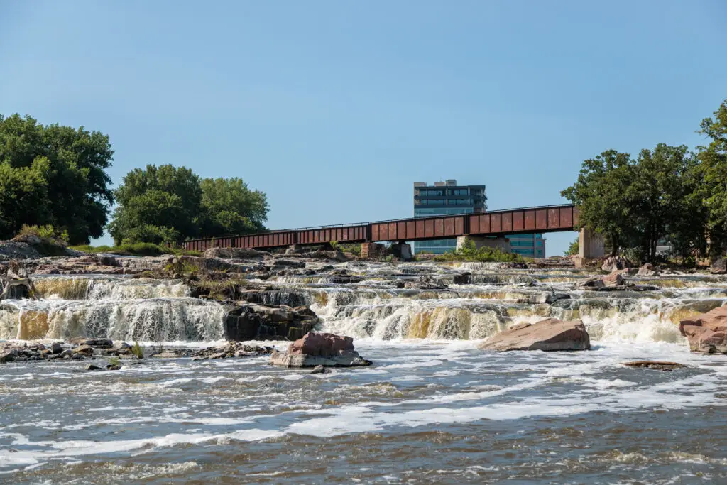 A river with small waterfalls flows under a rusty bridge, with trees and a building in the background.  Sioux Falls with Kids