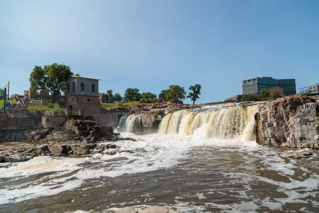 A waterfall cascades over rocky terrain with an old building on the left and trees in the background under a clear blue sky.  Sioux Falls with Kids