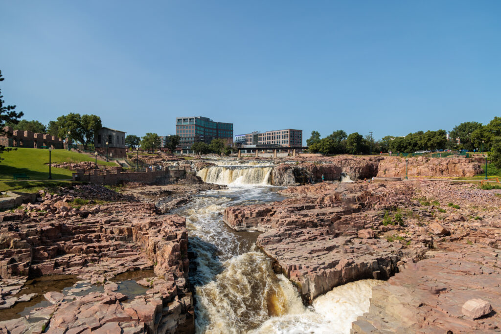 Wide view of Sioux Falls with cascading waterfalls, surrounded by rocky terrain, green spaces, and buildings in the background under a clear blue sky. Sioux Falls with Kids