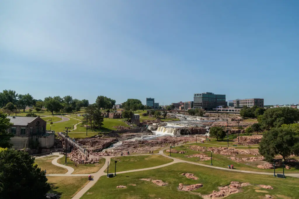 View of a city park with a waterfall, surrounded by paths, green lawns, and trees. Buildings are visible in the background under a clear blue sky.  Sioux Falls with Kids