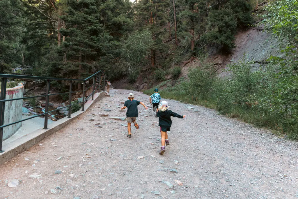 Three children run joyfully on a gravel path through a forested area, surrounded by trees and a metal railing on the left.  2 days in Ouray with kids.