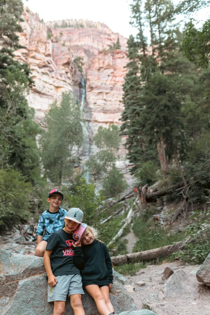 Three children sit on a rock in front of a waterfall surrounded by tall trees and rocky cliffs.  2 days in Ouray with kids.