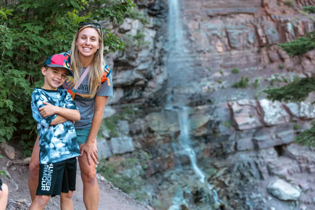 A woman and a child pose in front of a rocky waterfall. The woman is smiling, and the child is wearing a blue cap and tie-dye shirt. There are trees around them.  2 days in Ouray with kids.