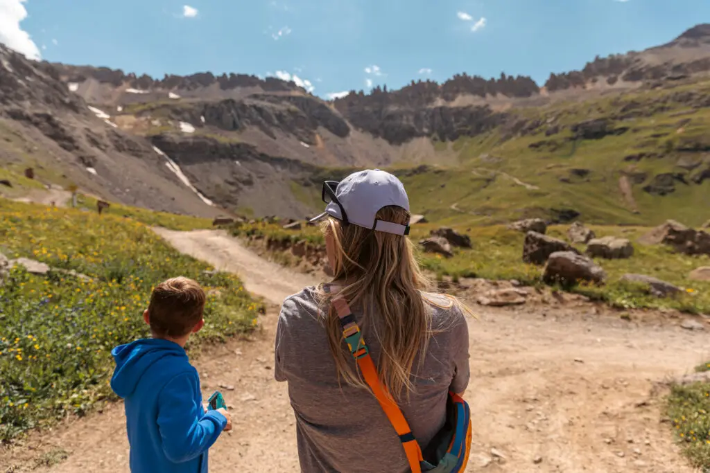 A woman and a child stand on a dirt path in a mountainous landscape under a clear sky. The woman is wearing a white cap and sunglasses.  2 days in Ouray with kids.