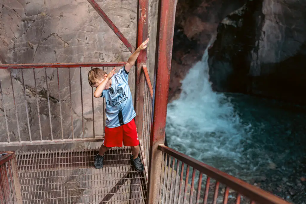 A child in a sports jersey and red shorts poses in a dab stance on a metal grate near a waterfall.  2 days in Ouray with kids.