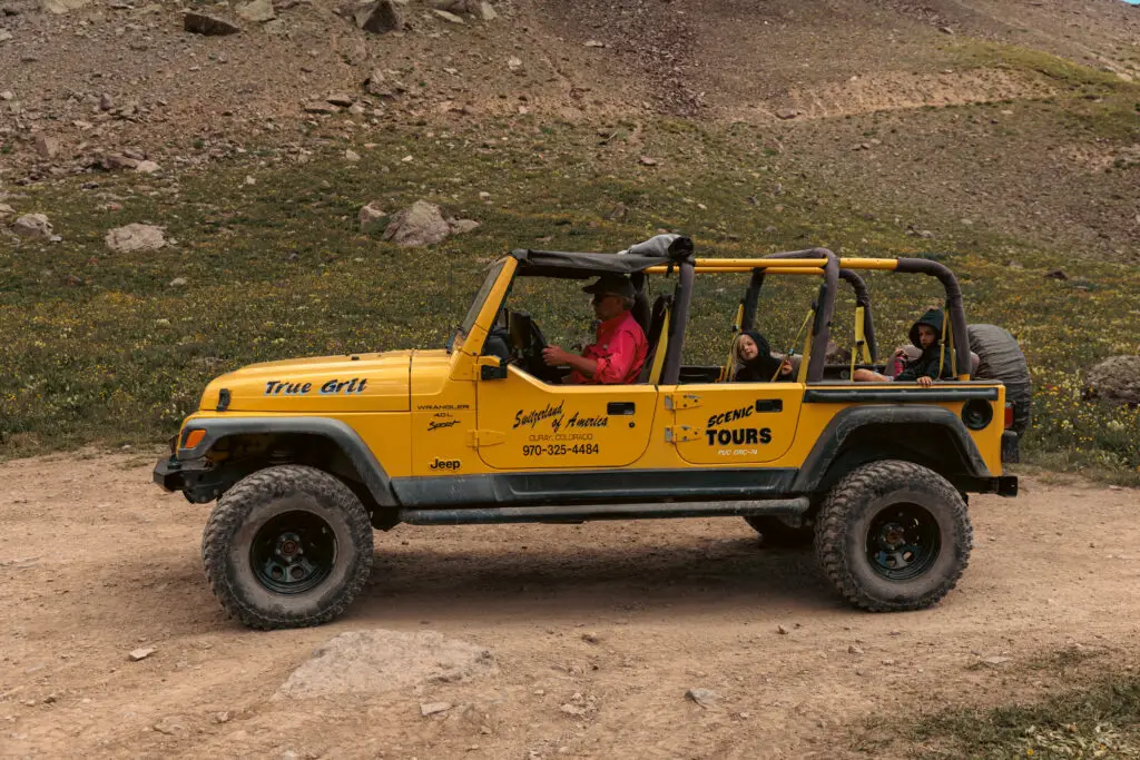 A yellow Jeep with "Scenic Tours" branding drives on a dirt road in a mountainous area. Several passengers are seated inside.  2 days in Ouray with kids.