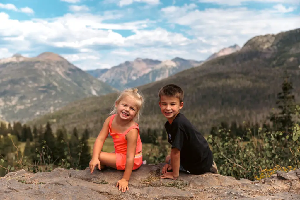 Two smiling children sit on a rock with mountains and trees in the background under a partly cloudy sky.  2 days in Ouray with kids.