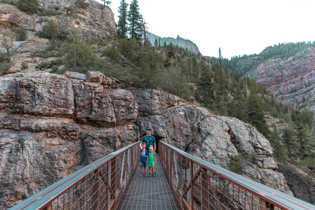 Three people stand on a narrow bridge surrounded by rocky cliffs and trees.  2 days in Ouray with kids.