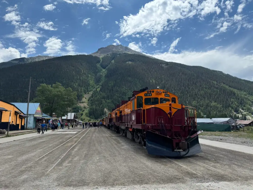 Red and yellow train on tracks in a small town, set against a backdrop of forested mountains under a partly cloudy sky.  Afternoon in Silverton with Kids.