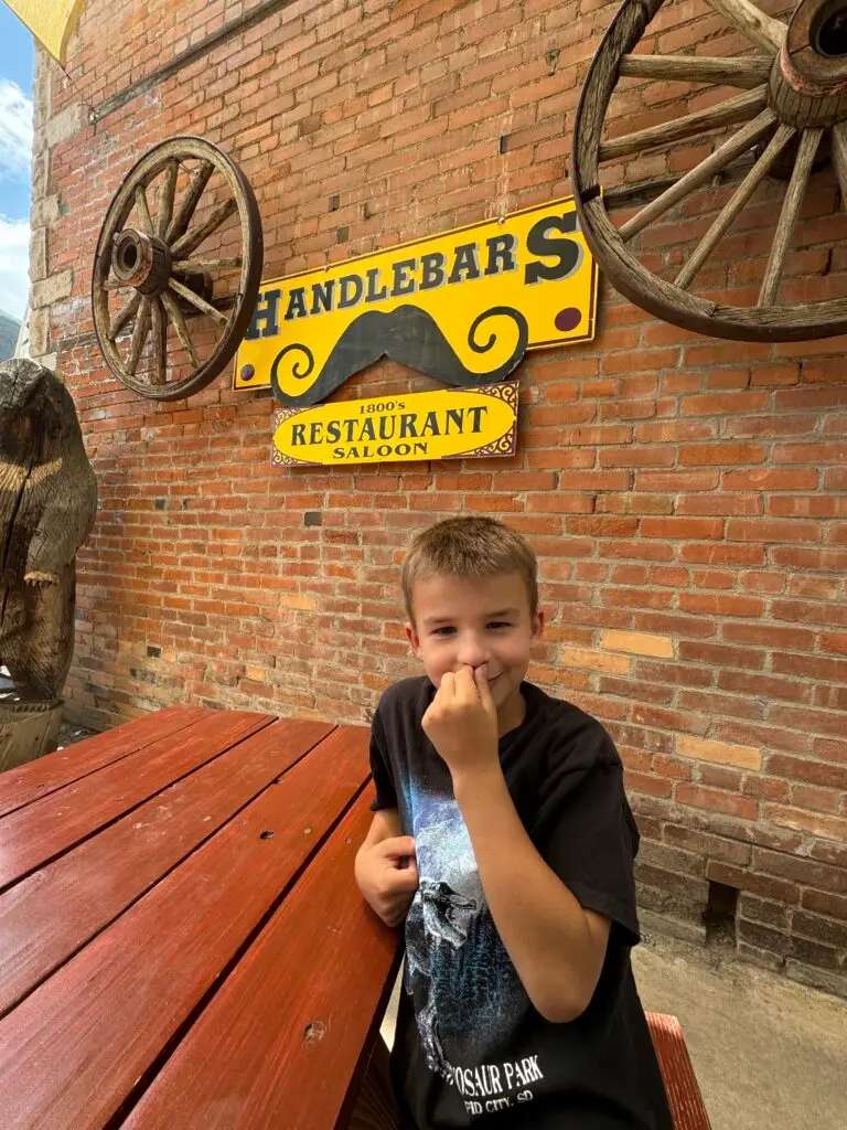 A boy sits at a wooden picnic table in front of a brick wall with a sign reading "Handlebars Restaurant Saloon.  Afternoon in Silverton with Kids.