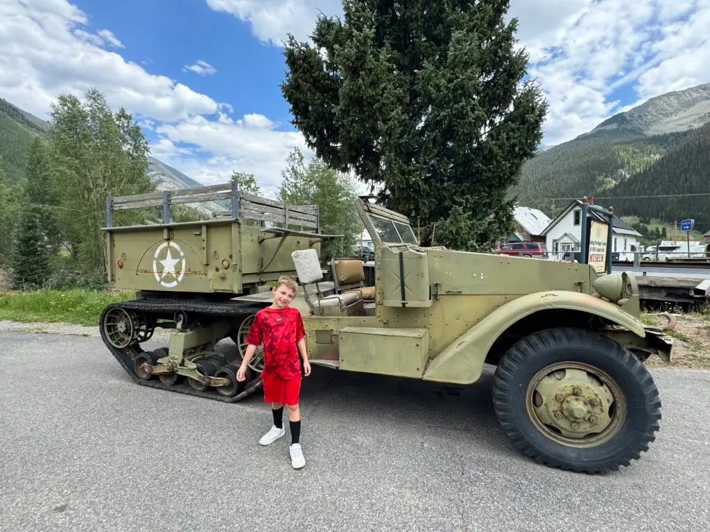 A child in a red outfit stands in front of an old military half-track vehicle on a road with mountains in the background.  Afternoon in Silverton with Kids.