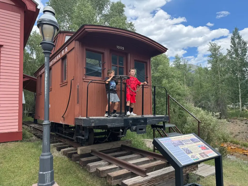 Two children stand on the platform of an old railroad car labeled "Colorado & Southern R.R." by a sign and lamp post, surrounded by trees and grass.  Afternoon in Silverton with Kids.