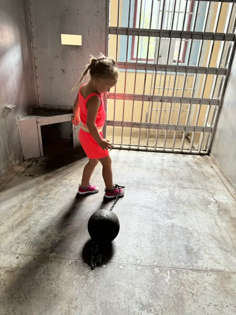 A young girl in a bright pink outfit stands in a cell, looking at a ball and chain on the floor.  Afternoon in Silverton with Kids.