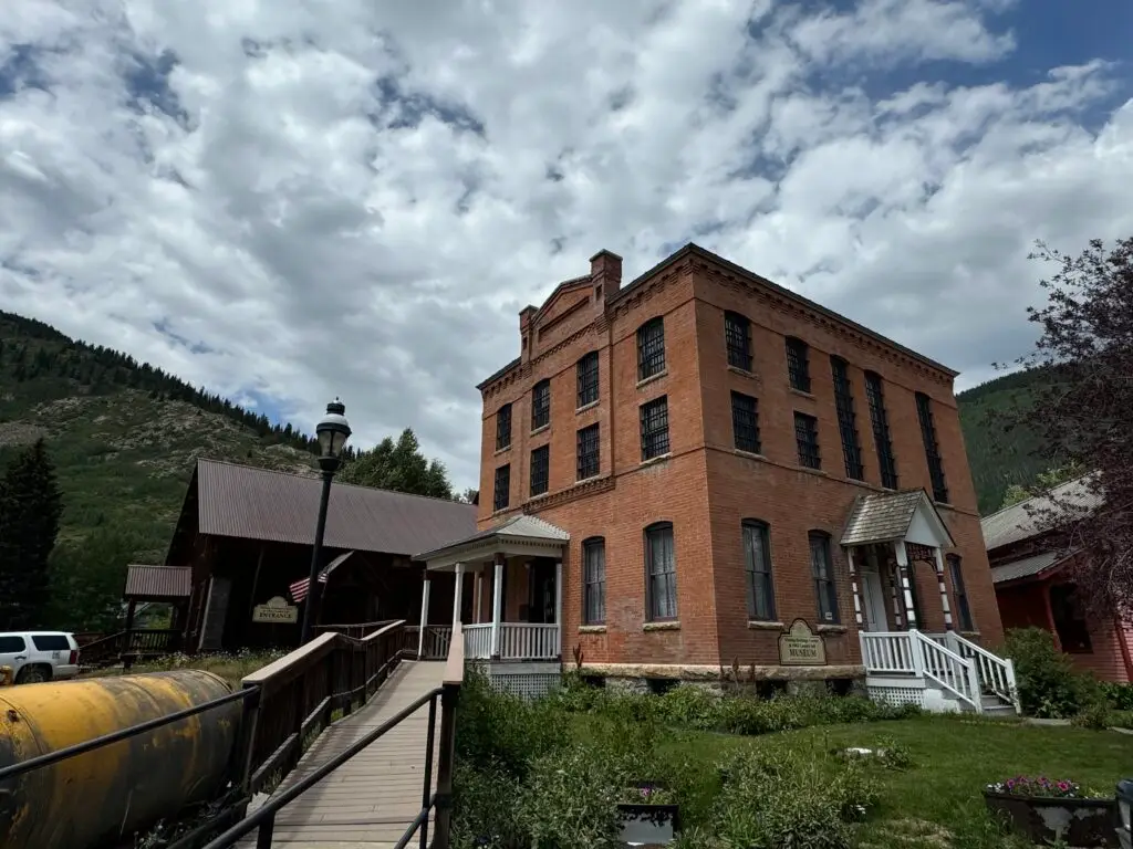 Historic brick building under a cloudy sky, with a walkway and small garden in front. Hills visible in the background.  Afternoon in Silverton with Kids.