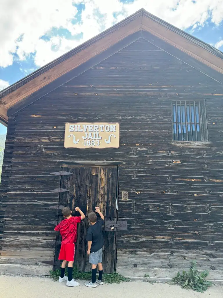 Two children stand in front of a weathered wooden building labeled "Silverton Jail 1883," looking at the door.  Silverton with kids.