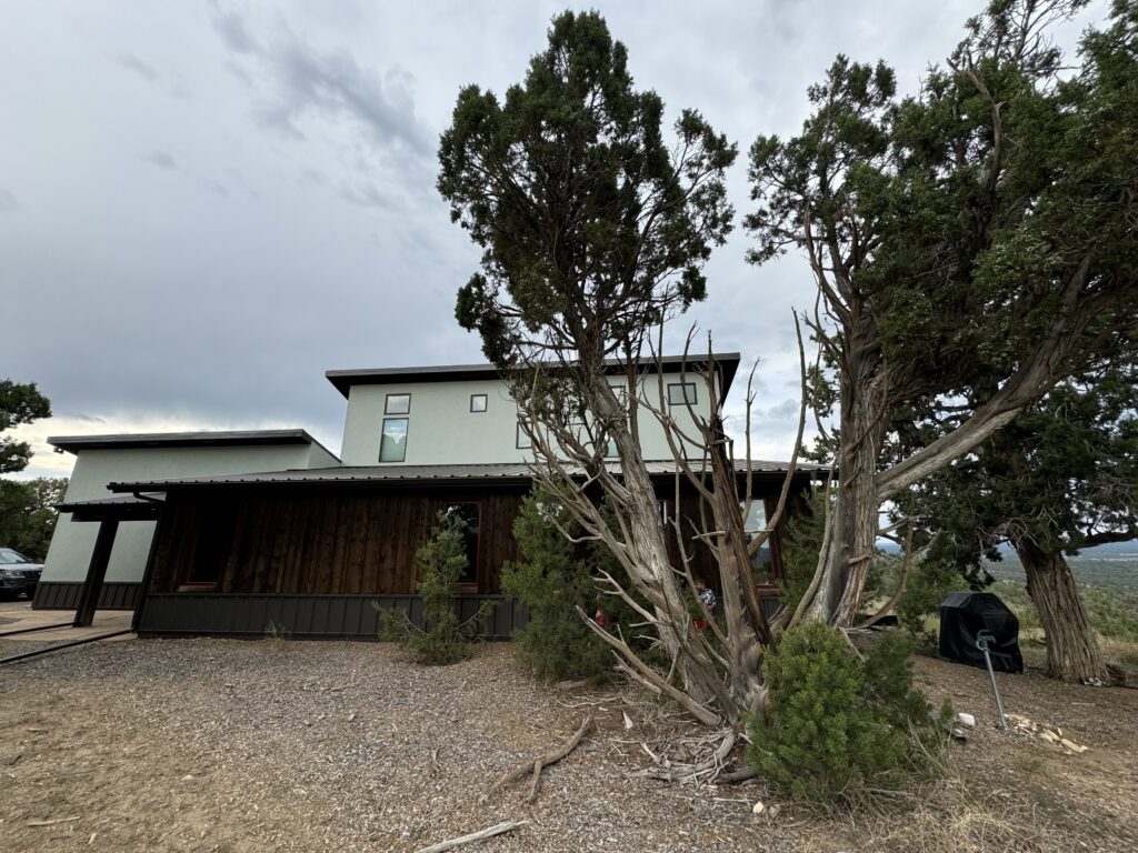 Modern two-story house with a flat roof and wooden siding, nestled like a Mesa Verde retreat, surrounded by trees and shrubs under a cloudy sky—perfect for families dreaming of cliffside adventures with the kids.  Mesa Verde with kids.