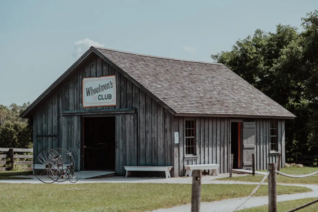 A rustic wooden building with a sign reading "Wheelmen's Club." A vintage bicycle is displayed outside. Surrounded by greenery.  Old World Wisconsin with Kids.  