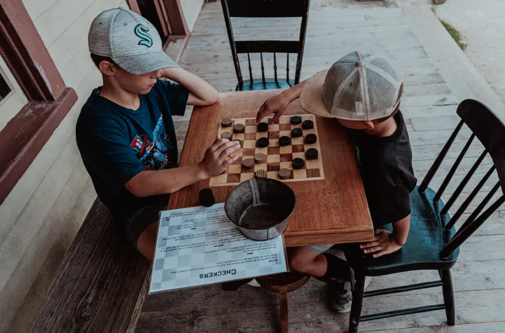 Two children play a game of checkers on a wooden table outdoors. A metal bucket and checkerboard instructions are on the table.  Old World Wisconsin with Kids.