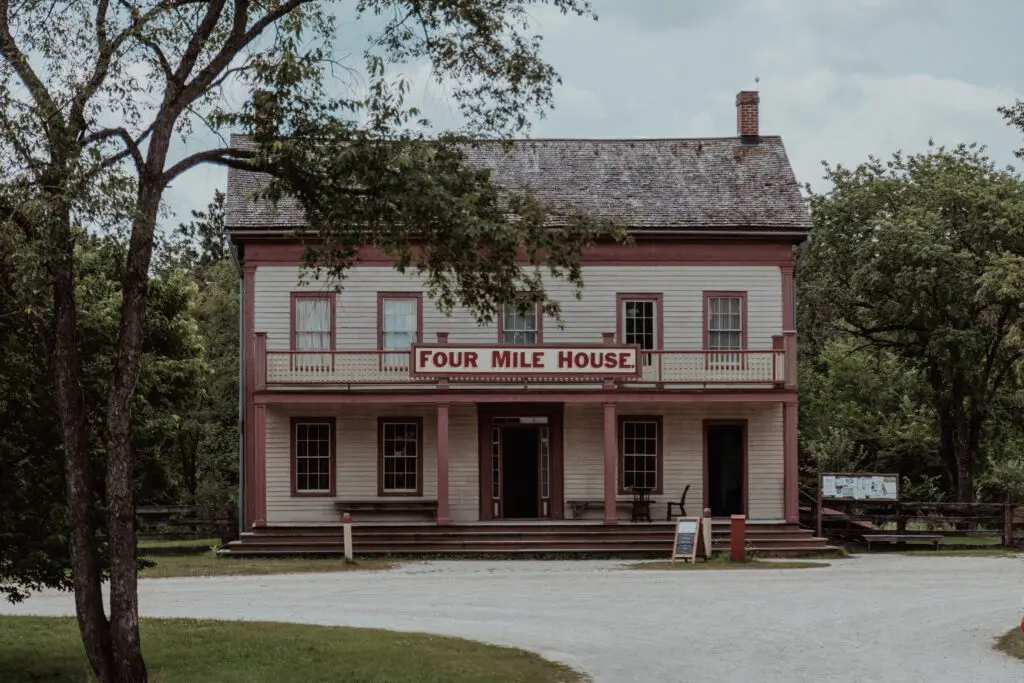 A historic wooden building with a sign reading "Four Mile House" sits surrounded by trees, with a gravel path in the foreground.  Old World Wisconsin with Kids.