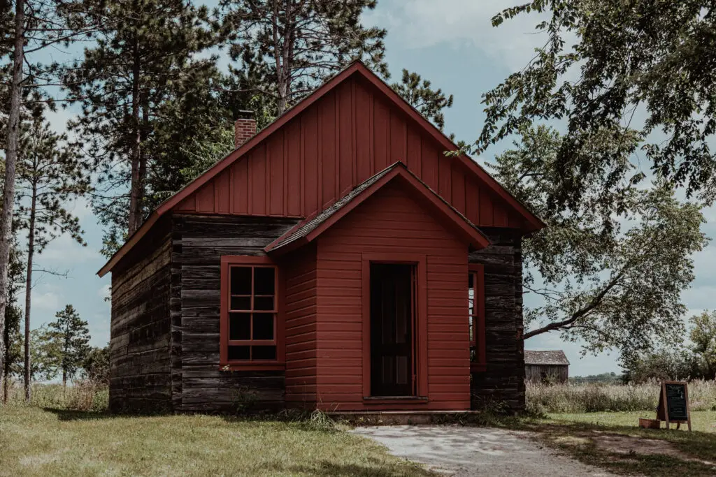 A small wooden cabin with red trim sits in a grassy area, surrounded by trees under a cloudy sky.  Old World Wisconsin with Kids.