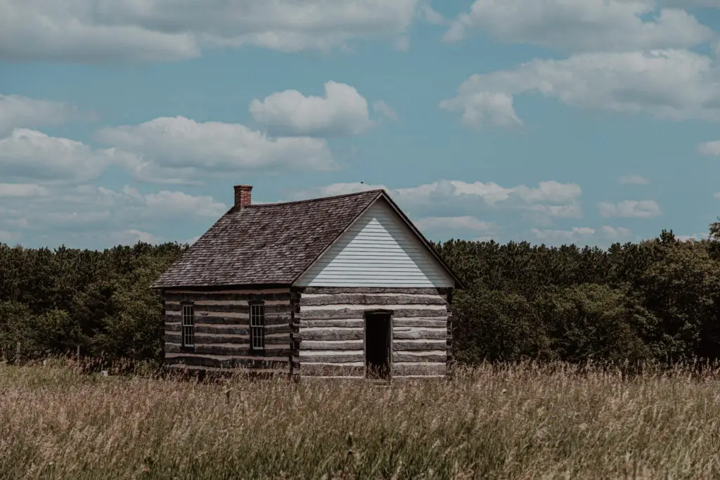 A small, rustic wooden cabin with a sloped roof stands in a grassy field under a cloudy sky, surrounded by trees.  Old World Wisconsin with Kids.