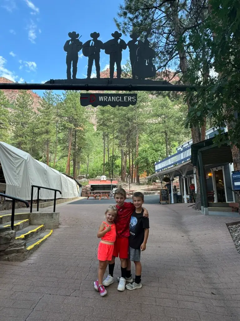 Three children pose for a photo under a sign with cowboy silhouettes in a park-like setting with trees and cabins.  Bar D Chuckwagon Suppers with kids