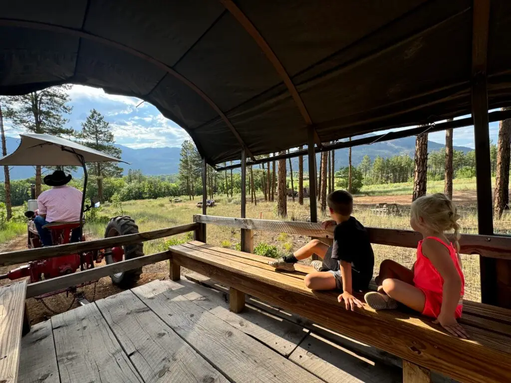 Two children sit on wooden benches in a covered wagon, looking out at a scenic view. A person on a tractor leads the ride through a wooded area with mountains in the background.  Bar D Chuckwagon Suppers with kids