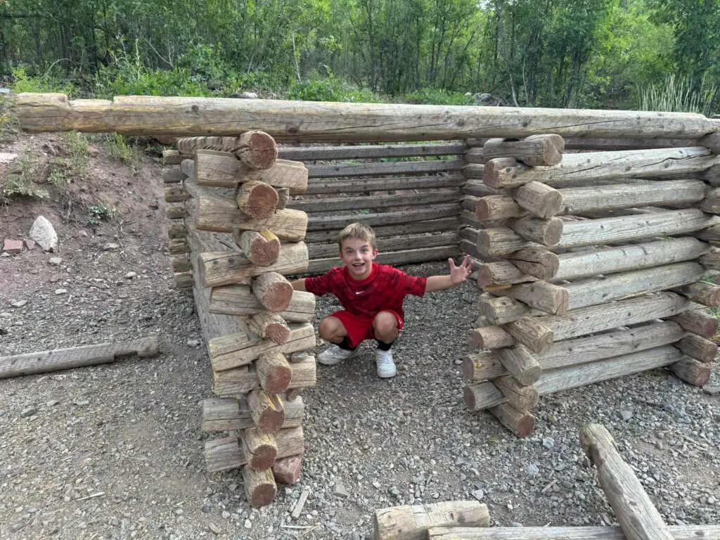 A child in a red shirt crouches and smiles inside a structure made of stacked wooden logs, set outdoors with trees in the background.  Bar D Chuckwagon Suppers with kids