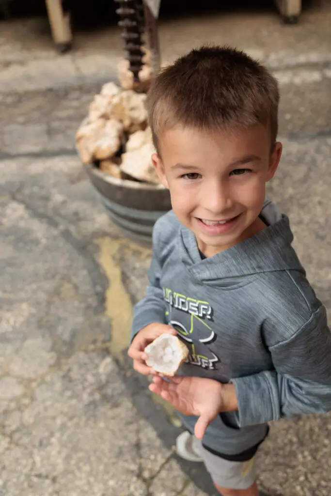 A young boy smiling while holding a rock, standing outdoors on a paved surface with a metal container full of rocks in the background.  Cave of the Mounds with kids.