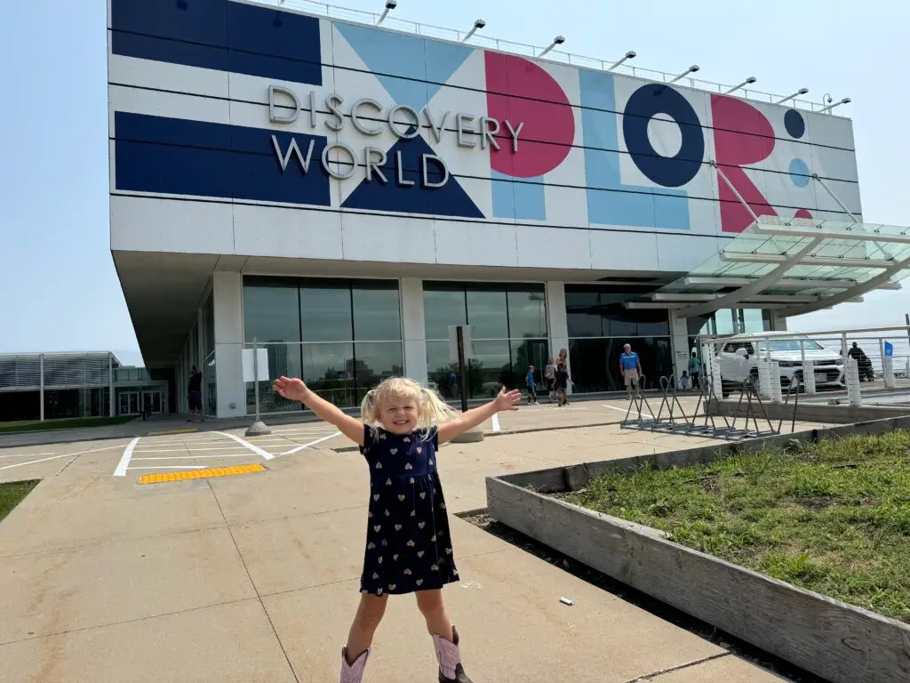 A young child with blonde hair and a blue dress stands excitedly outside the Discovery World building.