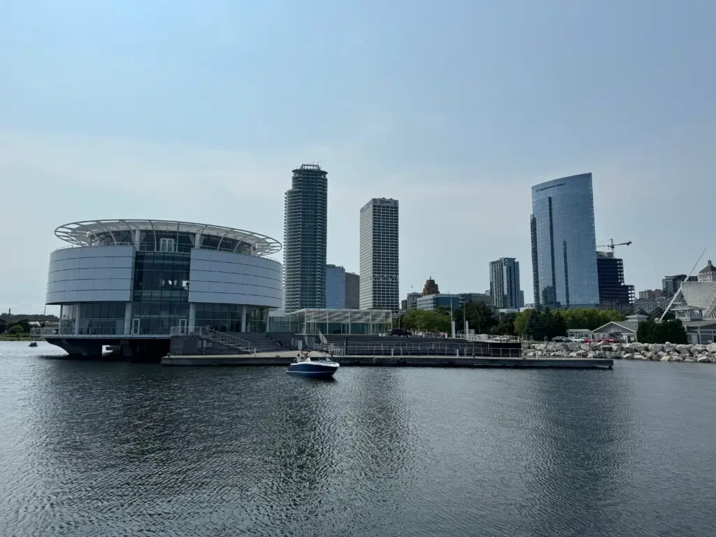 A waterfront scene with a circular building on the left and several modern skyscrapers in the background, a boat docked nearby under a clear sky.