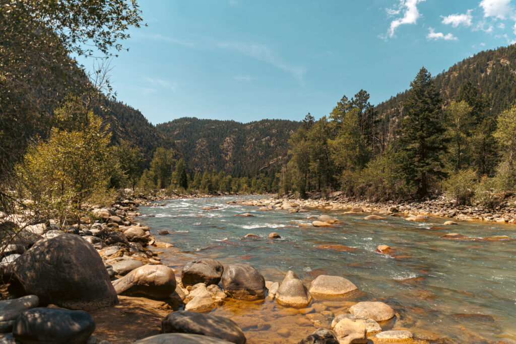 River flowing through a forested mountain landscape, with clear skies and rocky banks.  Durango train with kids.