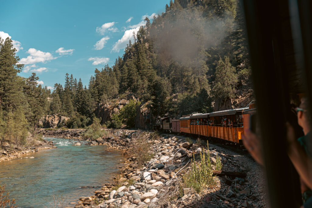 The Durango train travels through a forested mountainous area next to a rocky river under a clear sky, offering an unforgettable adventure for kids. Durango train with kids.