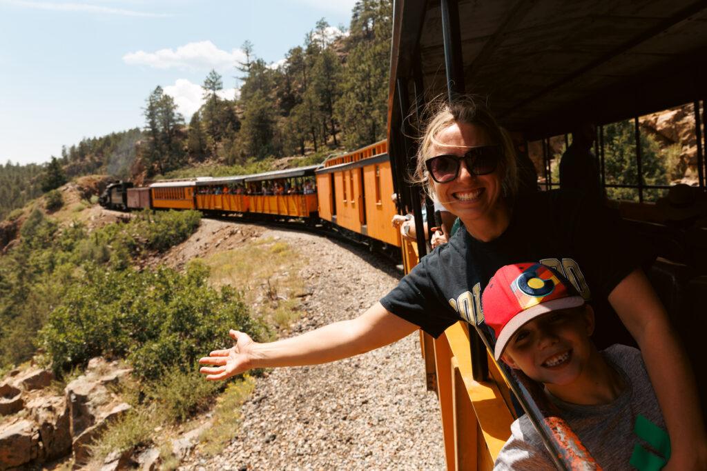 A woman and a child smile and gesture towards a scenic view from the open side of a moving train, with forested hills in the background.  Durango train with kids.