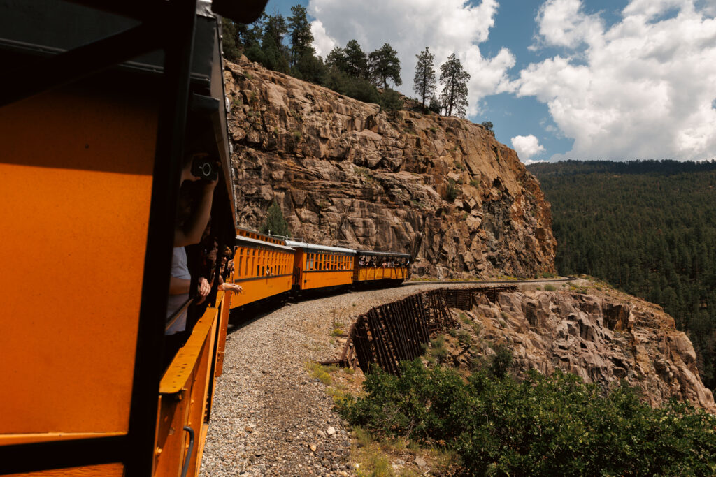 A yellow train travels on a curved track alongside a steep rocky cliff, surrounded by trees and under a partly cloudy sky. Passengers are visible leaning out with cameras.  Durango train with kids.