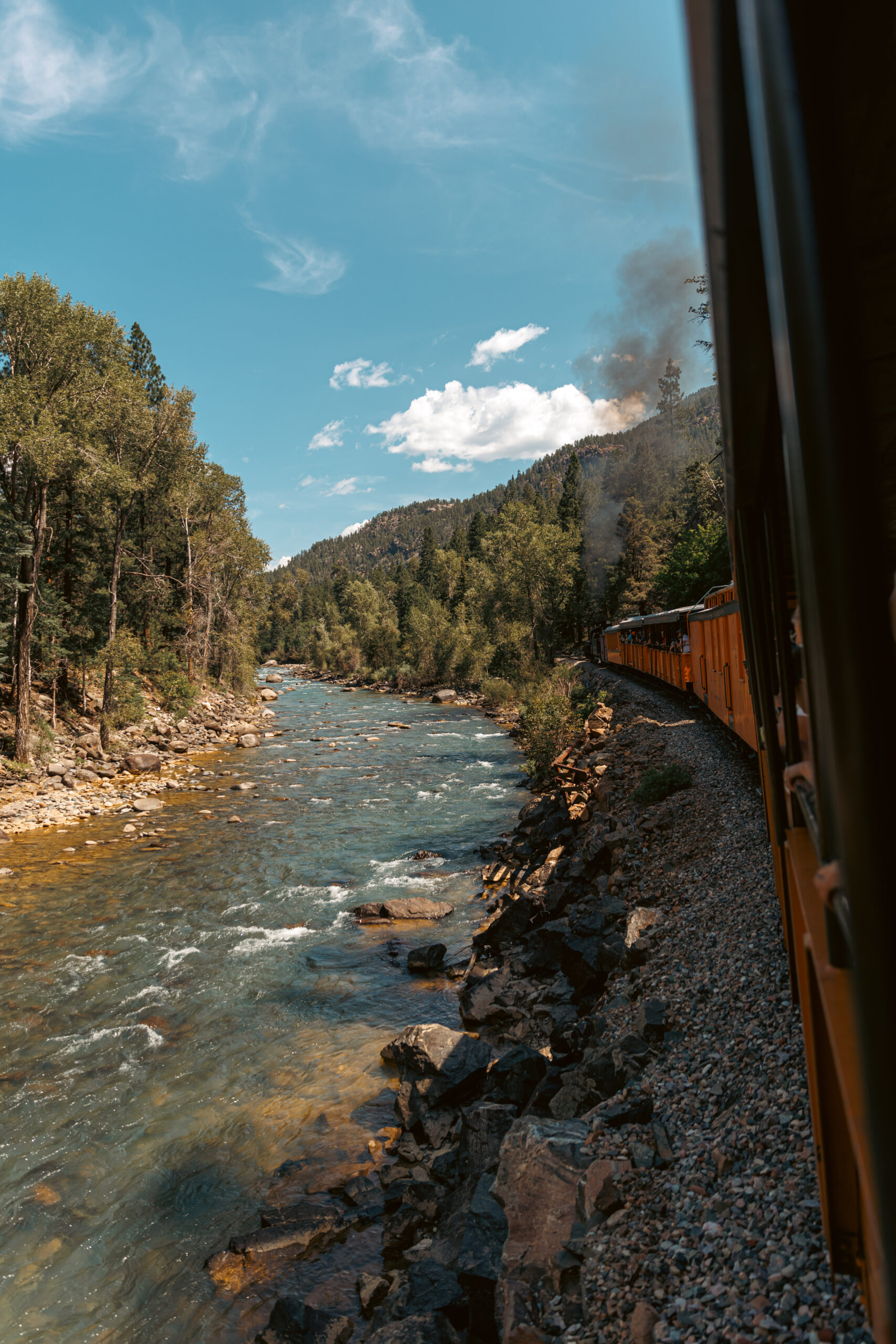 A Durango train chugs along a rocky riverbank, smoke billowing from its engine, capturing the imagination of kids. The picturesque landscape features trees and distant hills under a partly cloudy sky, making it an unforgettable journey for all aboard.  Durango train with kids.