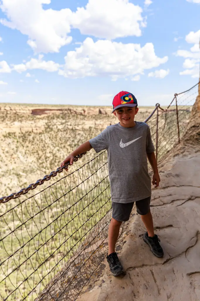 A young boy in a gray Nike shirt and red cap stands on a rocky path with a wire fence, overlooking the vast canyon landscape of Mesa Verde under a cloudy sky, capturing the spirit of adventure when exploring with kids. Mesa Verde with kids.